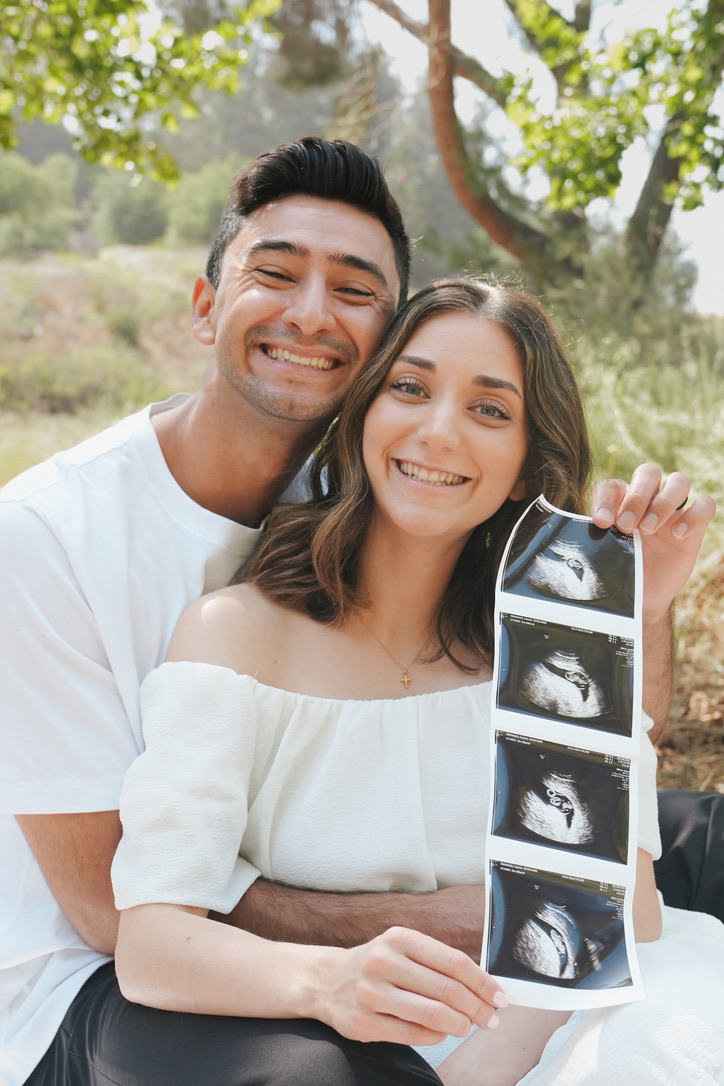 Beaming couple holding a strip of ultrasound photos beneath the trees