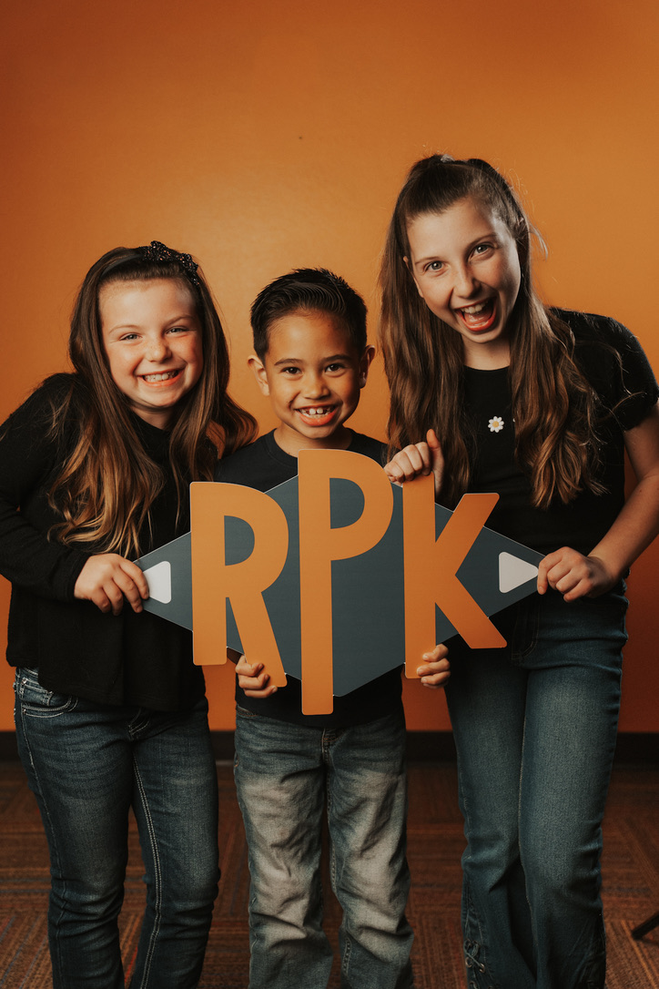 Three kids laughing while holding an RPK sign