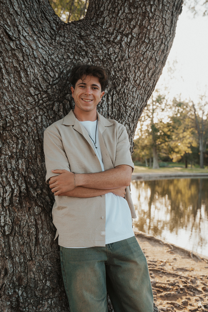 Arms-crossed portrait smiling beneath a towering tree by the water