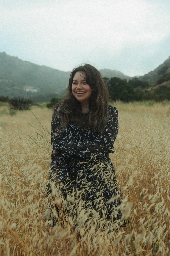 Woman in a floral dress smiling in a golden field beneath misty hills