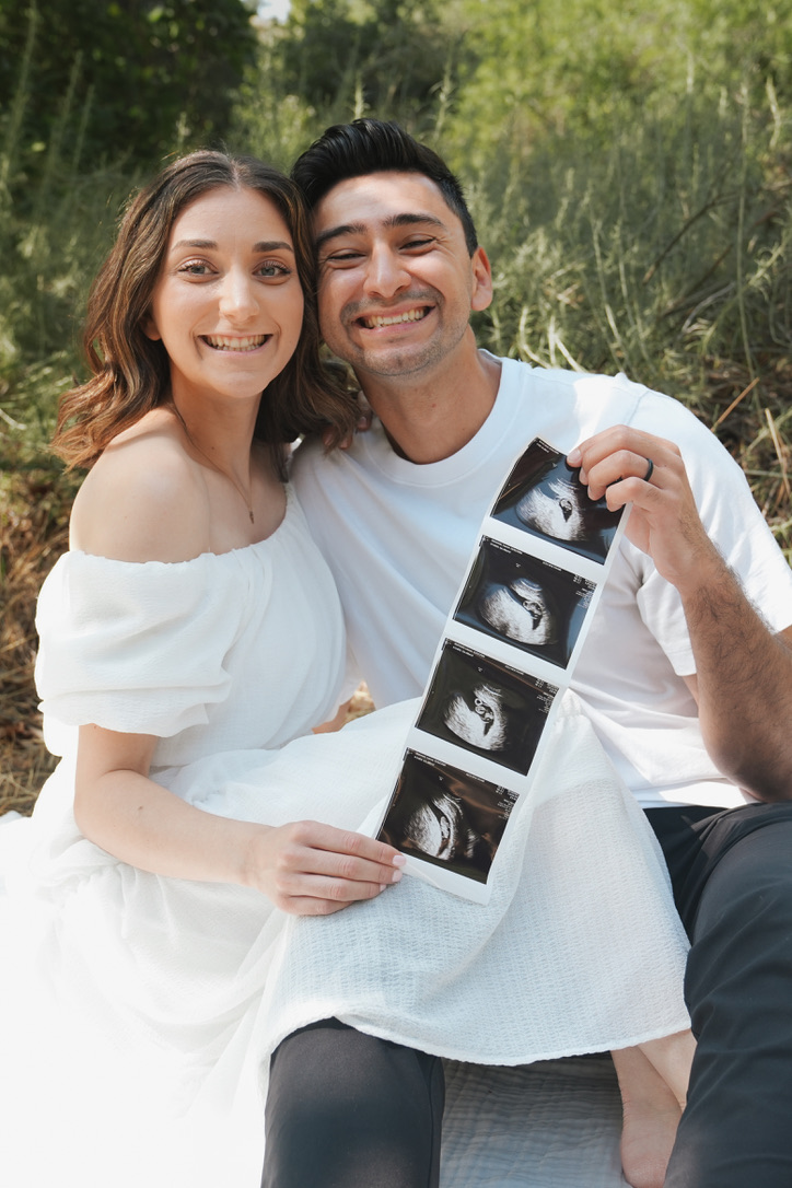 Seated couple smiling with a strip of ultrasound photos in tall grass