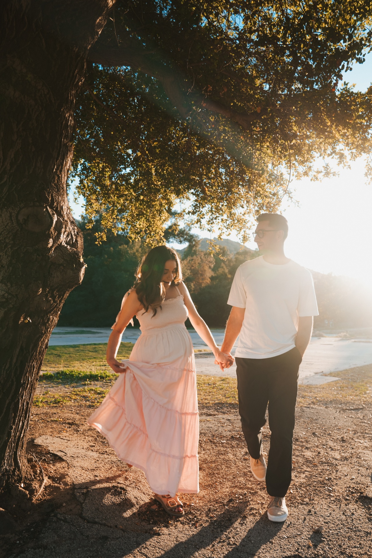 Expecting couple holding hands beneath a sunlit oak at golden hour