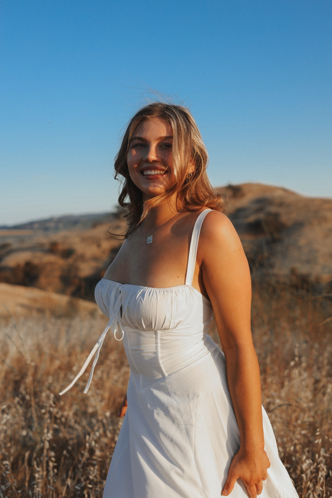 Sunlit smile in a white corset dress among golden grass and hills