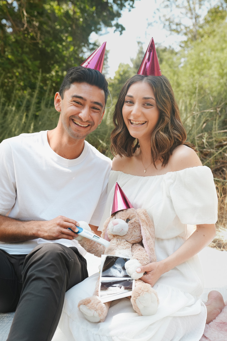 Couple in party hats celebrating a pregnancy announcement with a teddy bear